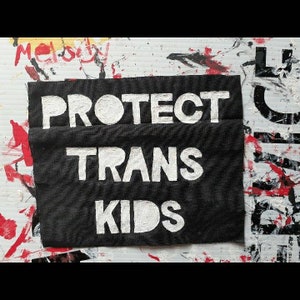 Könnte beinhalten: Ein schwarzes Stoffbanner mit den Worten "PROTECT TRANS KIDS" in großen, weißen Blockbuchstaben. Das Banner befindet sich vor einem Hintergrund aus Weiß und Grau mit roten und schwarzen Farbspritzern.