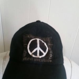 Pode incluir: Boné de beisebol preto com um emblema de símbolo de paz branco. O símbolo da paz é costurado em um patch quadrado. O boné tem uma aba curva e um botão na parte superior. Um acessório de moda.