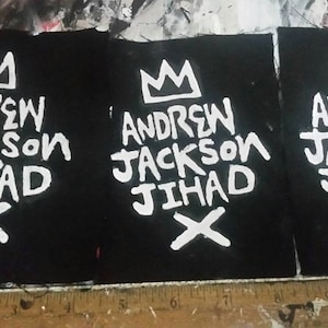 May include: Three black fabric patches with white text and a crown design. The text reads "ANDREW JACKSON JIHAD" with an "X" below. The patches are arranged side by side.