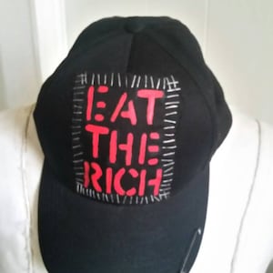Może przedstawiać: Czarna czapka bejsbolówka z czerwoną i białą naszywką z napisem "EAT THE RICH". Naszywka jest przyszyta do czapki białą nicią. Do czapki przymocowana jest srebrna agrafka.