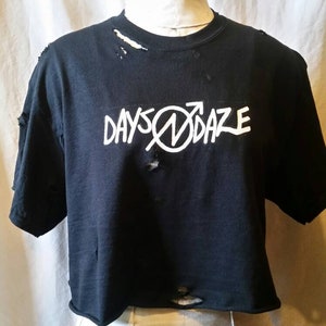 May include: Black, distressed crop top with the white text "DAYSØDAZE". The shirt has a round neckline and short sleeves. The fabric has several holes and tears, giving it a worn appearance. The shirt is displayed on a mannequin.