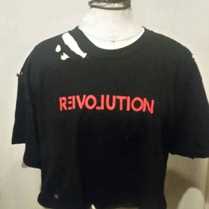May include: Black cropped t-shirt with the word "REIVOLUTION" in red letters. The shirt has distressed details, including holes and frayed edges. The shirt is displayed on a mannequin.