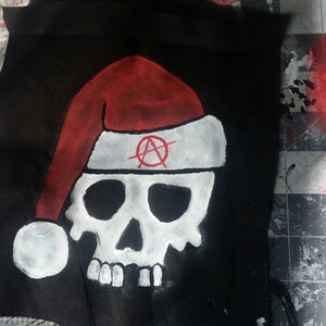 Puede incluir: Tela negra con una calavera blanca que lleva un gorro de Papá Noel rojo con borde blanco. El gorro tiene un símbolo de anarquía rojo. La calavera está pintada de blanco. El fondo es una superficie de trabajo desordenada.