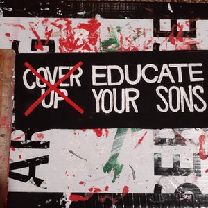 以下が含まれることがあります： 「EDUCATE YOUR SONS」と白い大文字で書かれた黒いバナーがある、ミクストメディアのアートワーク。赤い「X」が「COVER UP」の文字を消しています。背景は、赤と緑のペンキの飛沫がある白と黒のコラージュです。