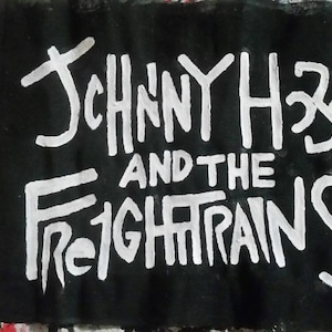 Pode incluir: Patch de tecido preto com a inscrição branca "JOHNNY H33" acima de "AND THE FREIGHTRAINS". O fundo é uma mistura de respingos de tinta vermelha, branca e preta.