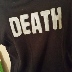 Könnte beinhalten: Schwarzes T-Shirt mit dem Wort "DEATH" in großen, weißen Blockbuchstaben aufgedruckt. Das T-Shirt wird über einem weißen Rollkragen getragen. Das Bild ist eine Nahaufnahme des T-Shirts.