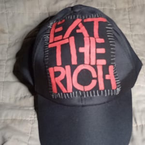Op de afbeelding: Zwarte baseballpet met een gebogen klep. De voorkant heeft een patch met de rode tekst "EAT THE RICH". De patch is omrand met witte stiksels.