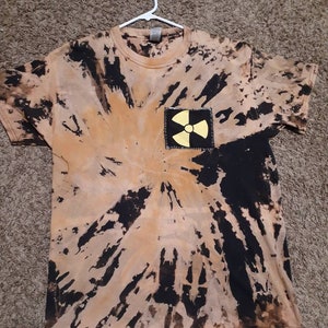 Könnte beinhalten: Kurzärmeliges T-Shirt mit Batikmuster in Beige- und Schwarztönen. Ein quadratischer Aufnäher mit einem gelben Strahlungssymbol ist auf der Vorderseite aufgenäht. Das T-Shirt hängt an einem weißen Kleiderbügel.