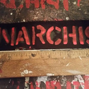 May include: A black rectangular patch with the word "ANARCHIST" in red, stencil-style lettering. A wooden ruler is placed below the patch. The patch is approximately 6 inches long.
