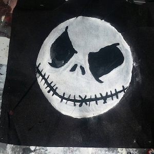 Könnte beinhalten: Ein schwarzer Stoff mit einem weiß gemalten Bild von Jack Skellingtons Gesicht aus The Nightmare Before Christmas. Das Gesicht hat große schwarze Augenhöhlen, einen genähten Mund und einen weißen ovalen Kopf.