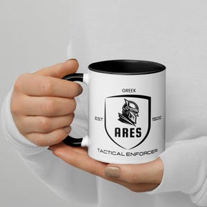 Puede incluir: Una taza de cerámica blanca con un borde y un asa negros. La taza presenta un gráfico en blanco y negro de un casco de caballero con el texto "GREEK EST 1500 ARES TACTICAL ENFORCER".