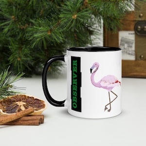 Könnte beinhalten: Weißer Keramikbecher mit schwarzem Innenraum und Griff. Der Becher zeigt eine rosa Flamingo-Illustration und das Wort "OBSERVER" in Grün. Der Becher hat eine Standardgröße für Kaffee oder Tee.