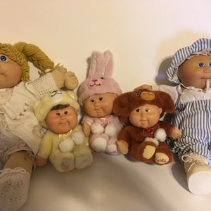 Puede incluir: Cinco muñecas Cabbage Patch Kids, cada una con un disfraz diferente. Las muñecas sonríen y tienen las mejillas sonrosadas. Las muñecas están sentadas sobre una superficie blanca.