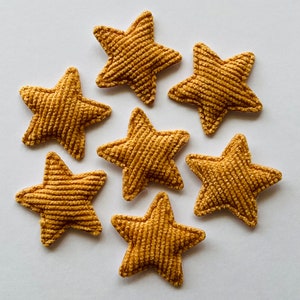 May include: Seven golden-yellow, corduroy-textured star-shaped appliques are arranged on a white surface. Each star has five points and a ribbed texture, creating a tactile and visually appealing design. These decorative stars are ideal for crafts and embellishments.