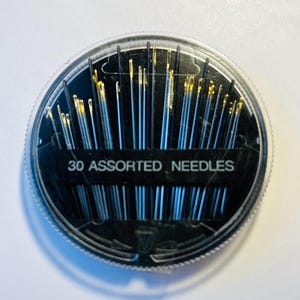 May include: A clear plastic round container with a black lid containing 30 assorted needles. The lid has the text "30 ASSORTED NEEDLES" printed on it.
