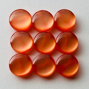 Può includere: Nove cabochon rotondi in vetro arancione disposti in una griglia 3x3. Ogni cabochon ha una superficie lucida e bombata, che riflette la luce. Il colore è un arancione caldo e traslucido, con una leggera variazione di tonalità. Sono su sfondo bianco.