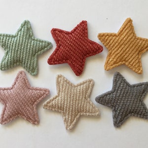 May include: Six star-shaped fabric patches in various colours, including green, red, yellow, pink, beige, and grey. The stars are made of a textured fabric with a corduroy-like texture.