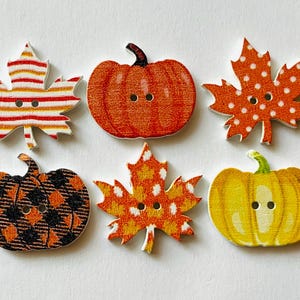 May include: Six decorative buttons shaped like autumn leaves and pumpkins. The set includes a striped leaf, a solid orange pumpkin, a polka-dot leaf, a black and orange plaid pumpkin, a mottled orange leaf, and a yellow pumpkin, each with two holes.