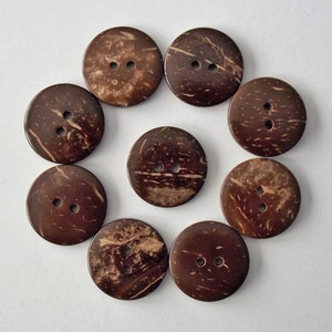 May include: A collection of ten round, dark brown coconut shell buttons with two holes each. The buttons have a natural, textured appearance with lighter brown streaks, arranged in a circular pattern against a white background. Ideal for sewing and craft projects.