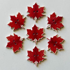 May include: Seven red maple leaf-shaped wooden buttons, each with two holes, arranged in a circle. The buttons have a distressed appearance with white edges, set against a white background. Suitable for sewing and craft projects.