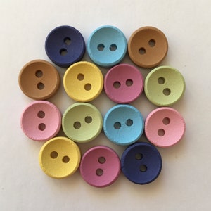 Könnte beinhalten: Sortiment von 12 bunten Holzknöpfen mit jeweils zwei Löchern. Die Knöpfe sind in Blautönen, Grün, Gelb, Braun und Rosa.