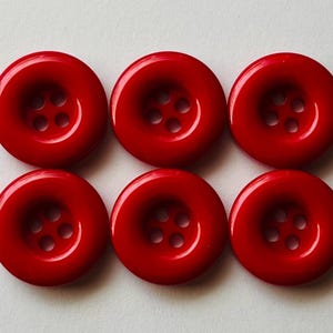 Peut inclure: Un ensemble de six boutons circulaires rouge vif disposés en deux rangées de trois. Chaque bouton présente une finition lisse et brillante ainsi qu'un centre en creux doté de quatre petits trous régulièrement espacés pour le passage du fil.