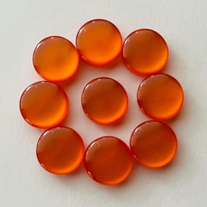 May include: Eight circular, smooth, polished carnelian gemstones arranged in a circular pattern. Each stone features a vibrant, translucent orange-red hue with a glossy finish, reflecting light on their rounded, convex surfaces.