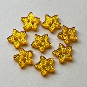 May include: A collection of eight star-shaped buttons in a vibrant yellow colour with a glittery finish. Each button features two small holes in the centre for attachment. The stars are arranged in a circular pattern, showcasing their uniform shape.