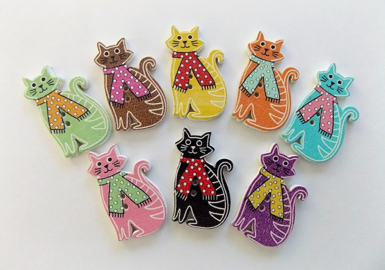 May include: A collection of nine wooden cat-shaped buttons in various colors, including green, brown, yellow, orange, pink, black, blue, and purple. Each cat wears a scarf with a polka dot pattern. The buttons have two holes for sewing.