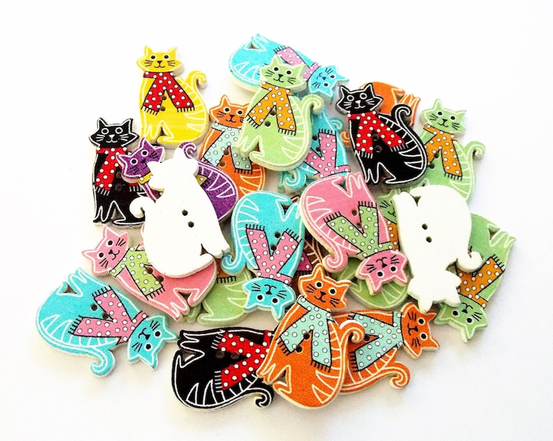May include: A collection of colorful wooden cat-shaped buttons. The buttons feature various colors, including yellow, black, orange, and green, with patterned scarves and polka dots. Some buttons have two holes for sewing.