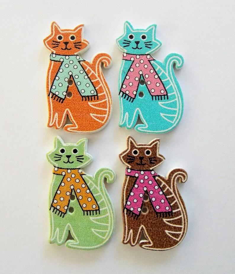 May include: Four wooden cat-shaped buttons in orange, turquoise, green, and brown. Each cat wears a scarf with polka dots. The buttons have a white outline and are suitable for sewing or crafting projects.