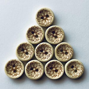 May include: A collection of nine circular light-colored wooden buttons arranged in a triangular formation. Each button features a two-hole center and a dark brown laser-engraved pattern of stylized leaves radiating from the middle to the edge.