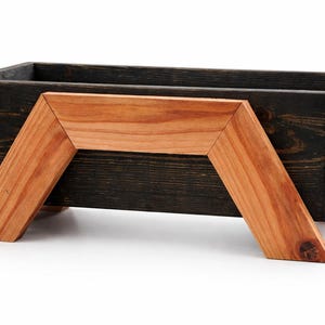 Może przedstawiać: Prostokątna drewniana skrzynka na rośliny z ciemno bejcowanym zewnętrzem i naturalnym drewnianym wspornikiem. Donica ma ciemno bejcowane wnętrze i jest podparta dwoma kątowymi, jasnymi drewnianymi nogami.