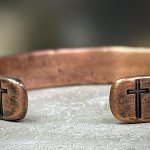 May include: A copper cuff bracelet with a hammered texture. The open ends of the bracelet each feature a carved cross. The bracelet has a rustic, antique appearance.