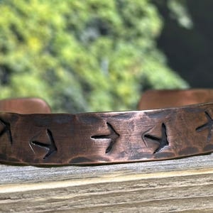 Puede incluir: Una pulsera de puño de cobre con una textura martillada. La pulsera presenta una serie de símbolos de flecha grabados. La joya rústica se exhibe sobre una superficie de madera con un fondo verde borroso.