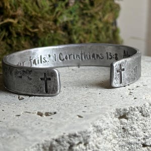 May include: A silver-toned cuff bracelet with a distressed finish. The bracelet features an engraved cross on each end and the text "1 Corinthians 13:8" along the top. The bracelet is resting on a textured surface.