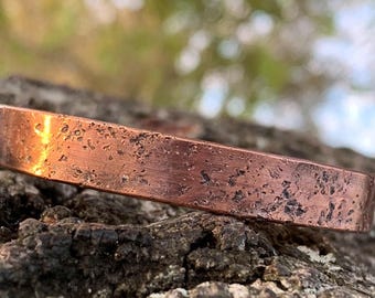 Puede incluir: Una pulsera de cobre con una superficie texturizada. La pulsera tiene un aspecto rústico y desgastado, con manchas oscuras y un color marrón rojizo cálido. La pulsera se encuentra sobre un fondo natural borroso.