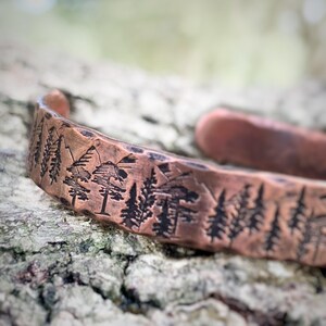 May include: A copper cuff bracelet with a nature-inspired design. The bracelet features an etched landscape of trees and mountains. The metal has a hammered texture and a warm, reddish-brown color.