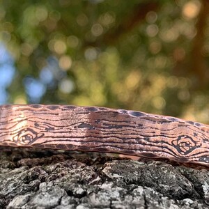 May include: A copper-colored metal bracelet with a wood grain texture. The bracelet is resting on a textured, dark gray surface. The bracelet has a rectangular shape and is slightly curved.