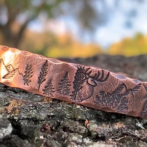 May include: Copper cuff bracelet with a hammered texture. The bracelet features an engraved forest scene with mountains, trees, and a deer. The cuff is approximately 0.75 inches wide.