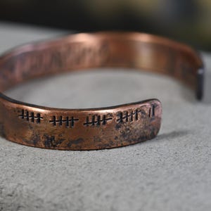 Puede incluir: Una pulsera de puño de cobre con un acabado envejecido. La pulsera presenta marcas de conteo grabadas en la parte delantera. El diseño abierto permite un uso fácil. La pulsera es de color marrón rojizo cálido.