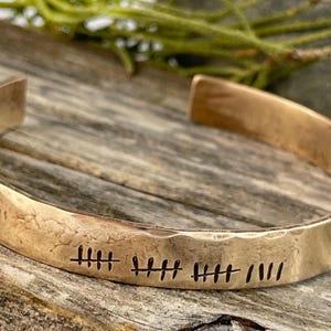 Puede incluir: Una pulsera de puño de cobre martillado con un diseño abierto. La pulsera presenta líneas verticales grabadas. La pulsera se muestra sobre una superficie de madera envejecida. Una joya única.