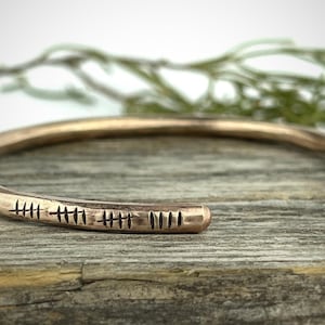 May include: A hammered copper cuff bracelet with etched markings. The open-ended bracelet has a rounded shape and a warm, metallic hue. The markings are evenly spaced and add a unique, handcrafted detail to the jewelry.