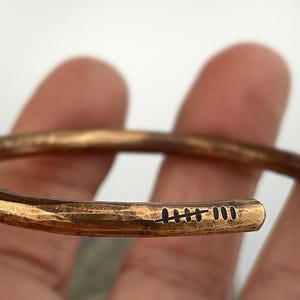 May include: A copper cuff bracelet with a hammered texture. The open end of the bracelet features a series of etched lines. The bracelet is held in a hand, showcasing its warm, metallic color and simple design.