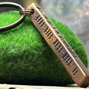 May include: A bronze-colored rectangular keychain pendant with etched markings and the phrase "The Journey of a Thousand Miles Begins with a Single Step." The pendant is attached to a metal keyring and rests on a bed of green moss.
