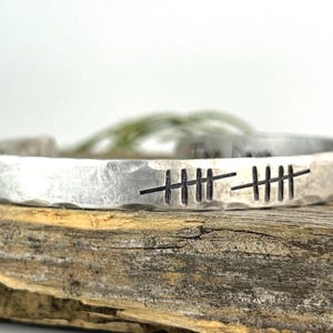 Puede incluir: Una pulsera de metal plateado con una textura martillada. La pulsera presenta marcas de conteo grabadas, probablemente representando un conteo o un hito. Se muestra sobre una superficie de madera.