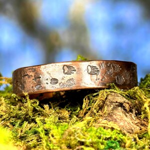 May include: A copper ring with stamped animal track designs. The ring features bear paw prints, deer tracks, and other symbols. The ring is set against a backdrop of green moss and a blurred blue and green background.