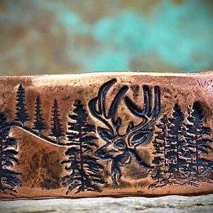 Puede incluir: Una pulsera de metal color cobre con una escena detallada de un bosque. El diseño presenta un ciervo con grandes astas, pinos y colinas. El fondo tiene un degradado turquesa y marrón.