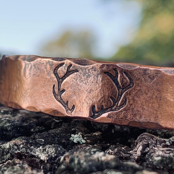 May include: Close-up of a copper-colored cuff bracelet with a hammered texture. The bracelet features a dark etched design of deer antlers. The bracelet is resting on a textured, dark surface.