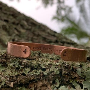 May include: A copper cuff bracelet with a hammered texture. The bracelet has the letter "A" stamped on one end and the letter "C" on the other. The text "sweet Bean" is engraved on the bracelet.